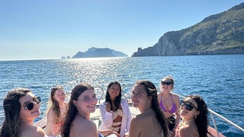 Group of six women in swimsuits on a boat under a bright sun with cliffs and sea in background.