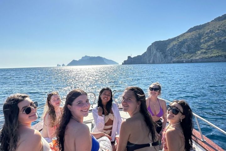 Group of six women in swimsuits on a boat under a bright sun with cliffs and sea in background.