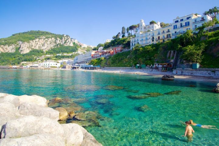 People swimming in clear water near rocky shore with hillside buildings.