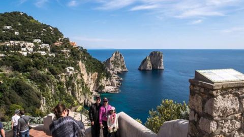 Tourists on a cliffside overlook with sea stacks in the distance and a clear blue sea and sky.