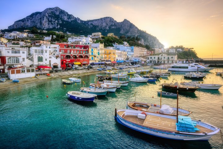Colorful coastal town with boats in harbor, mountains in background at sunset.