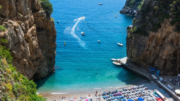 Small beach with umbrellas between cliffs, boats in blue sea.