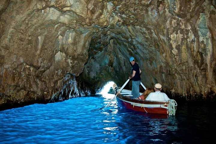 Small boat with people inside a rocky cave with blue water and light at the end of the tunnel.