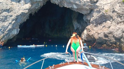 Woman on boat bow, people swim near rocky cave entrance in blue water.