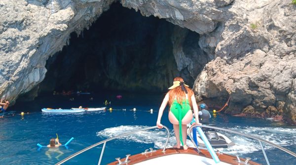 Woman on boat bow, people swim near rocky cave entrance in blue water.