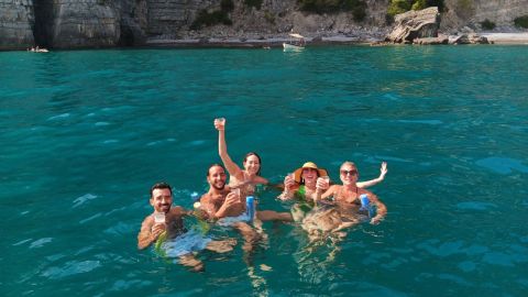 Five people floating in clear blue water, raising drinks near rocky cliffs and trees.