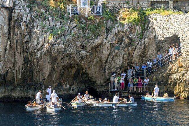 Tourists in small boats by a rocky cave entrance with people on a stone staircase.