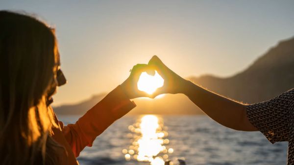 Two hands forming heart shape with sunset over water in background.
