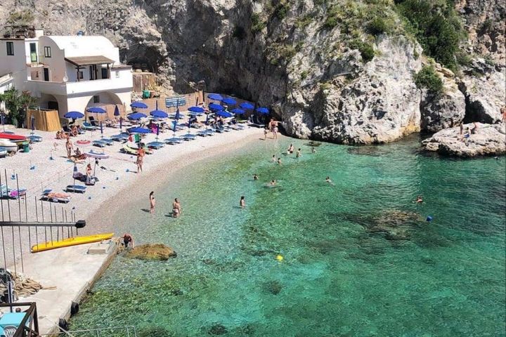 Beach with umbrellas, swimmers, boats in clear water, and rocky cliffs.