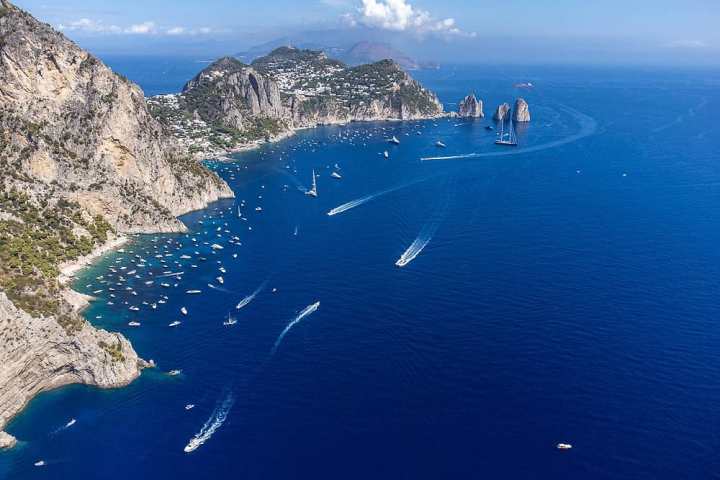 Aerial view of a rocky coastline with blue sea, boats, and distant cliffs under a clear sky.