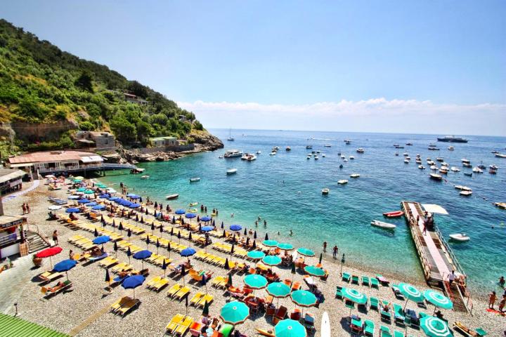 Pebble beach with colorful umbrellas and loungers, boats on the sea, and a nearby cliff.