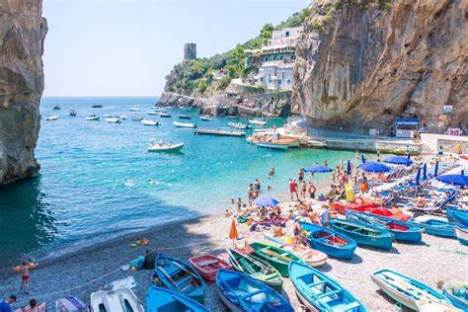 Beach scene with colorful boats, people sunbathing, rocky cliffs, and distant tower.