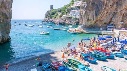 Beach scene with colorful boats, people sunbathing, rocky cliffs, and distant tower.