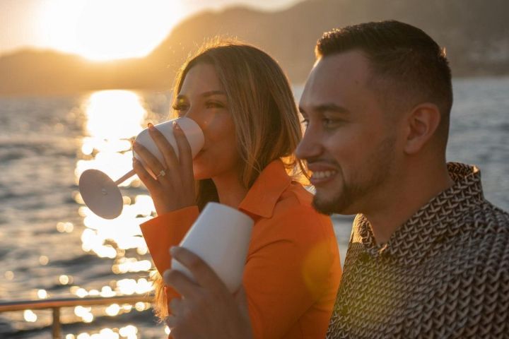 Couple on a boat at sunset, holding white glasses and smiling.