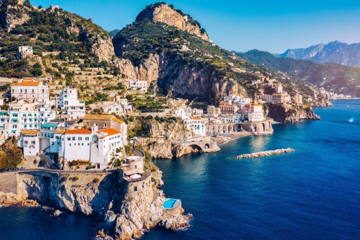 Scenic coastal village with colorful buildings on rocky cliffs, surrounded by blue sea and mountains.