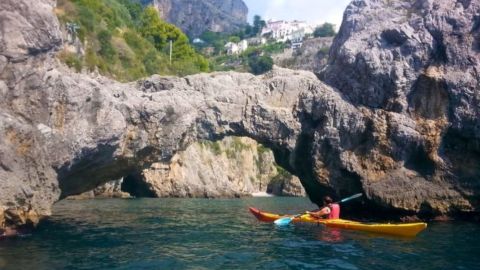Person kayaking under a natural rock arch near rocky cliffs and trees.