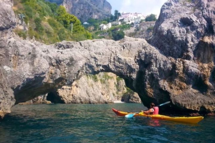 Person kayaking under a natural rock arch near rocky cliffs and trees.