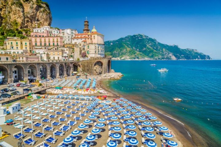 Beach with blue umbrellas, cliffside buildings, and boats on clear water.