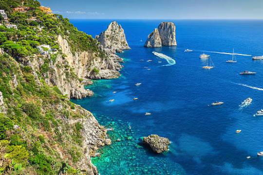 Coastal cliffs with boats in blue ocean and rocky formations at a distance under blue sky.