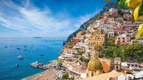 Colorful coastal town with hillside houses and boats in the sea under a clear blue sky, lemons in foreground.