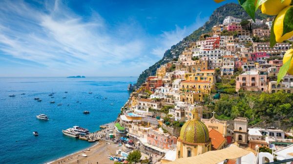 Colorful coastal town with hillside houses and boats in the sea under a clear blue sky, lemons in foreground.