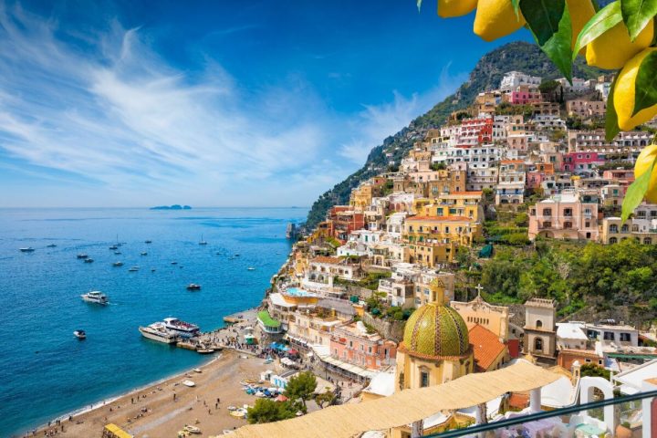 Colorful coastal town with hillside houses and boats in the sea under a clear blue sky, lemons in foreground.