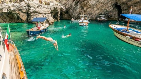 Person diving into turquoise water from a boat near rocky cliffs, with two other boats nearby.