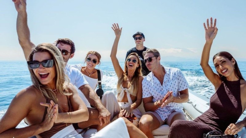 Group of people celebrating on a boat, smiling, with arms raised and ocean in background.